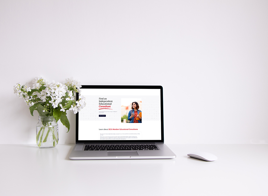 Laptop displaying a website for finding independent educational consultants, positioned on a minimalistic desk with a vase of white flowers and a computer mouse nearby.
